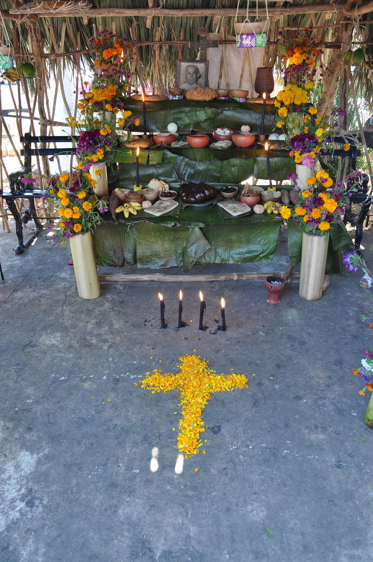 Altar under a thatched roof with marigolds, bowls of food, bread and a portrait, with four lit black candles and a cross of yellow petals on the ground in front