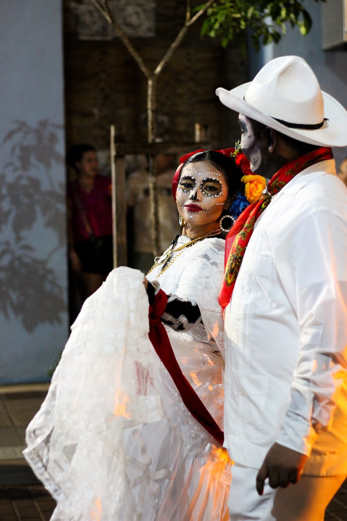 Une femme maquillée en catrina, robe de dentelle blanche et fleurs dans les cheveux, à côté d'un homme en blanc au chapeau blanc et foulard rouge, le visage peint