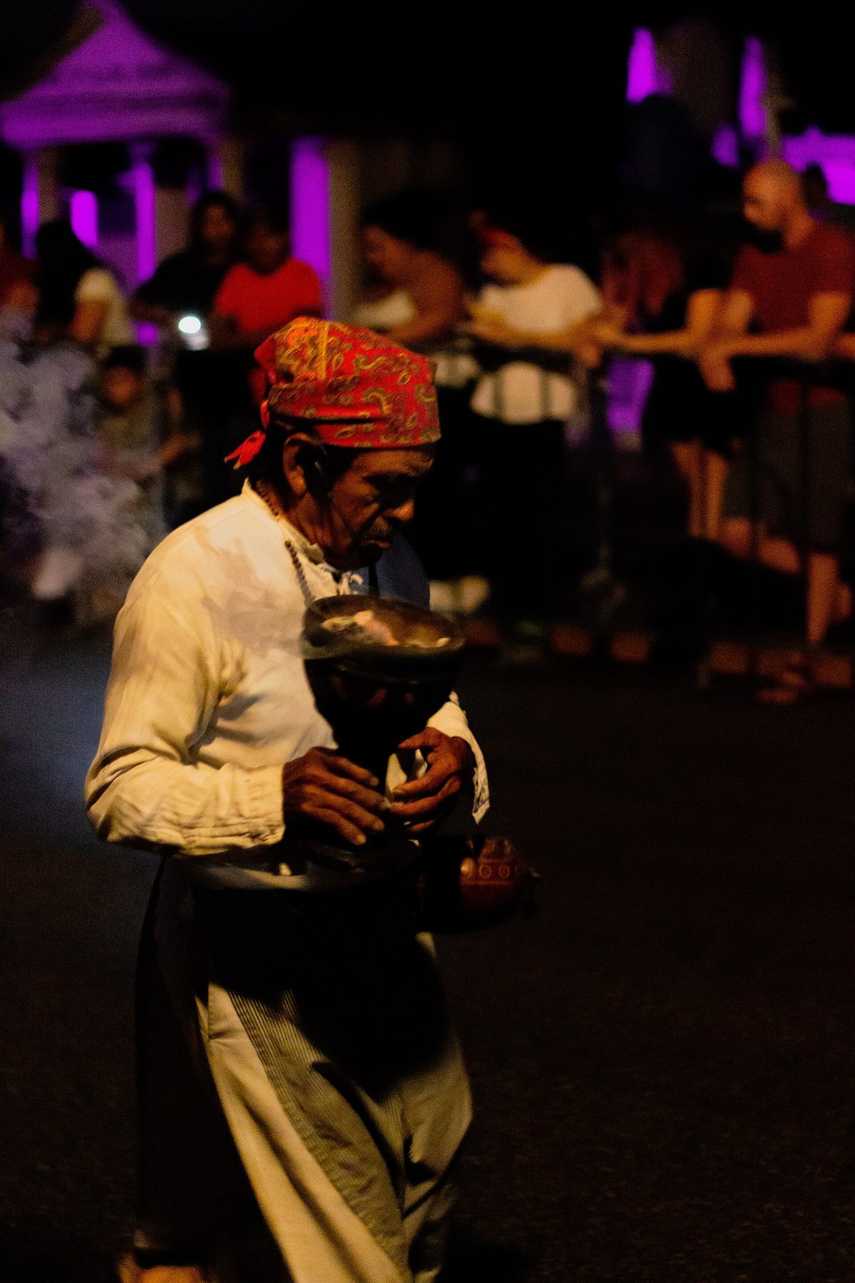 Un homme âgé vêtu de blanc, foulard rouge sur la tête, porte un encensoir fumant sur le parcours de la procession, la nuit
