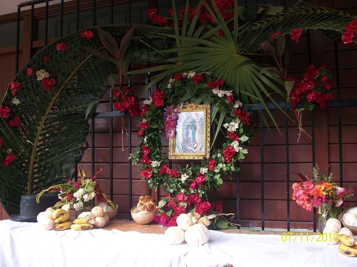 Autel familial sur une nappe blanche : image de la Vierge de Guadalupe dans une couronne de fleurs rouges, arche de palmes tressées, bananes, agrumes pelés et pain
