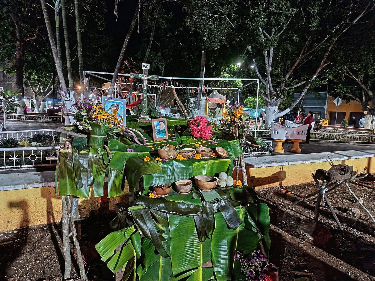 Autel dressé de nuit sur une structure de bois surélevée couverte de feuilles de bananier, avec une croix, une photo encadrée, des fleurs orange et des calebasses